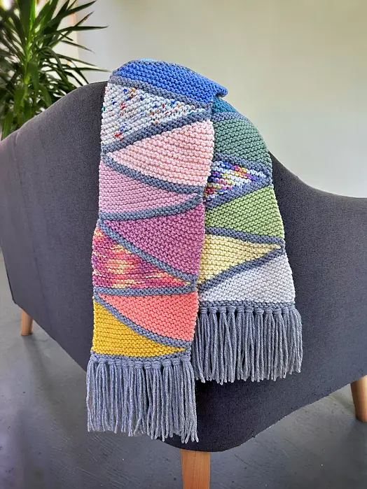 Free knitting pattern for a colorful scrap yarn scarf draped over the back of a gray sofa with gray fringe.